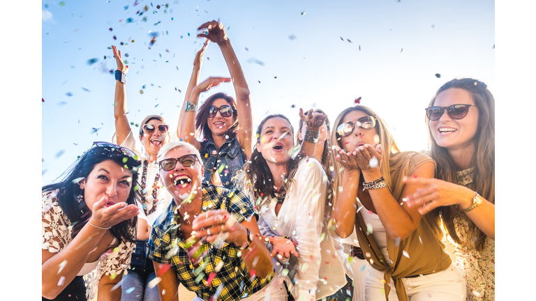 Cheerful group of happy people mixed ages generations women having fun all together during celebration party or carnival - view of fiends blowing. coloured confetti and laugh a lot in friendship