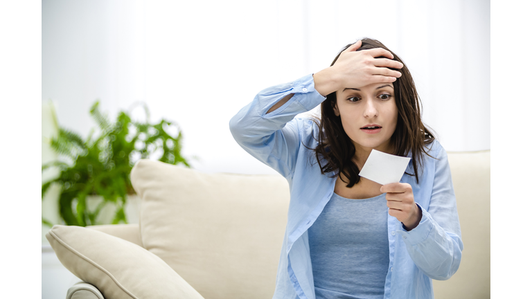 Confused and shocked woman is holding lottery ticket and looking at it, sitting on sofa. She is going to failure.
