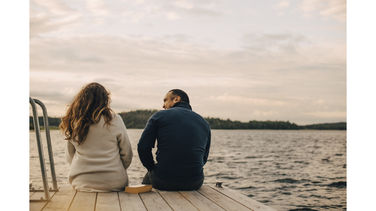 Rear view of couple talking while sitting on jetty by lake against sky