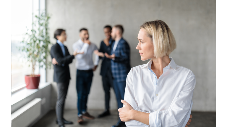 Male Coworkers Whispering Behind Back Of Unhappy Businesswoman In Office