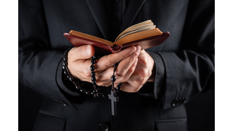 Hands of a christian priest dressed in black holding a crucifix and reading New Testament book.