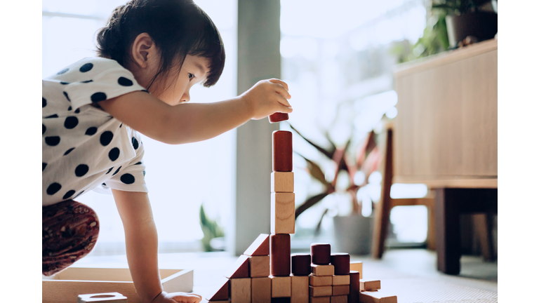 Creative little Asian girl crouching on the floor playing with wooden building blocks at home