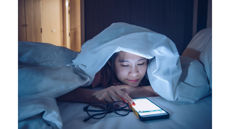 Portrait of woman hiding under the blanket, chatting and surfing on the Internet with smart phone late at night on bed.