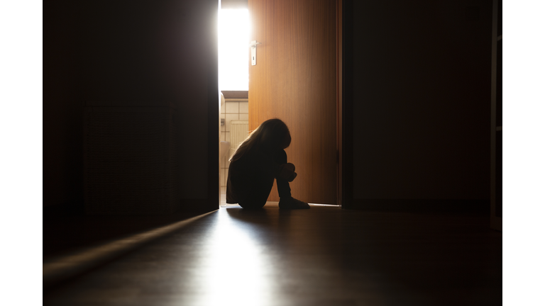 Despairing child sitting with head on knees in the dark frame of a doorway, backlit by a room behind flooded with daylight