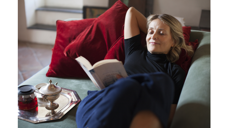 Beautiful mature woman relaxing on the sofa with cup of tea and a book