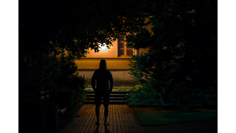 Hooded man standing on sidewalk at dark park in summer black night. Scary moment and gloomy atmosphere. Thief watching house. Back view.