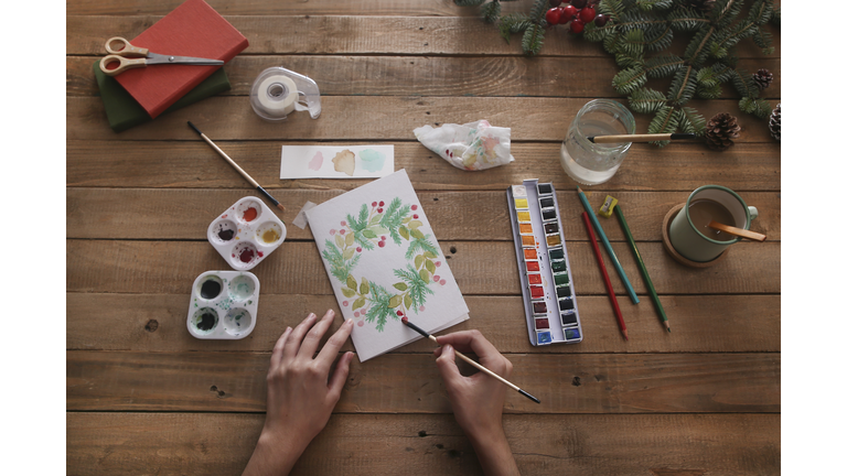 Young woman painting Christmas card with water colors, top view