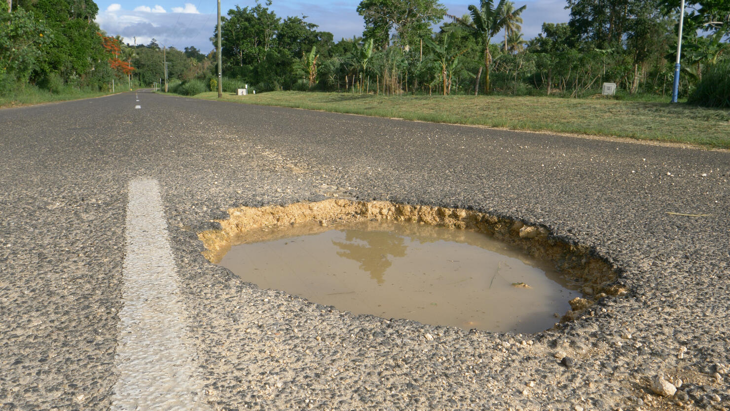 CLOSE UP: Tropical sun shines on large pothole in the middle of decaying road.