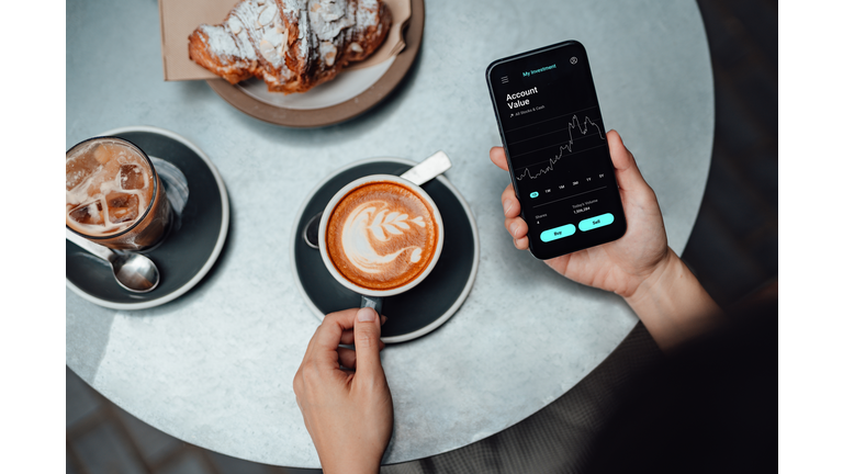 Young Woman Using Stock Trading App On Smart Phone At Cafe