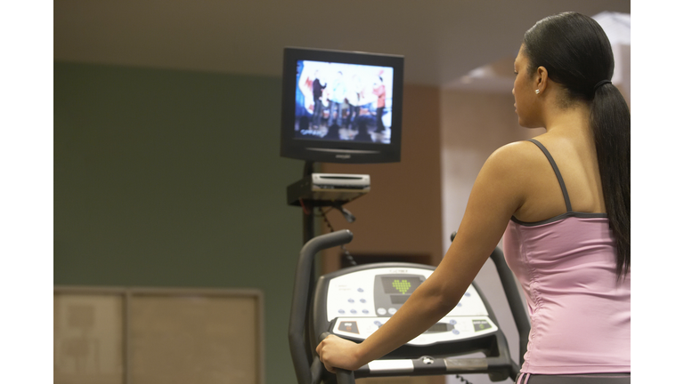 Young woman on treadmill, television in background, rear view