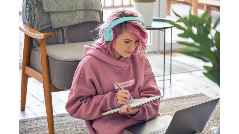 Focused hipster teen girl school college student pink hair wear headphones write notes watching webinar online video conference calling on laptop computer sit on floor working learning online at home.