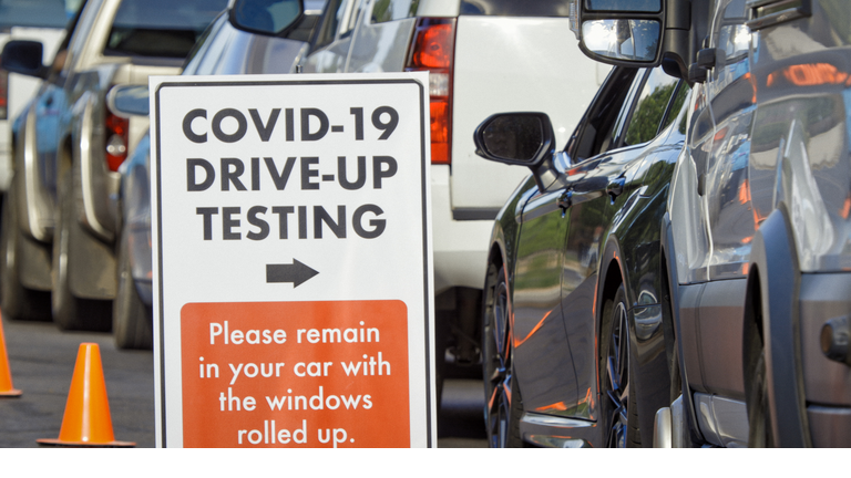 A "COVID-19 Drive-Up Testing" Sign Sits in the Foreground While Cars and Other Vehicles Wait in a Drive-Up (Drive Through) COVID-19 (Coronavirus) Testing Line Outside a Medical Clinic/Hospital Outdoors (Second Wave) in the Background