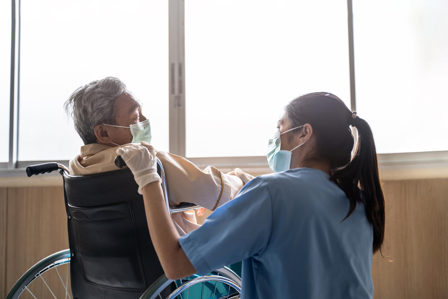 Asian nurse taking care of mature male patient sitting on wheelchair in hospital. Young woman and old man wearing surgical face mask for protection of covid 19 pandemic. Girl smile to elderly man.