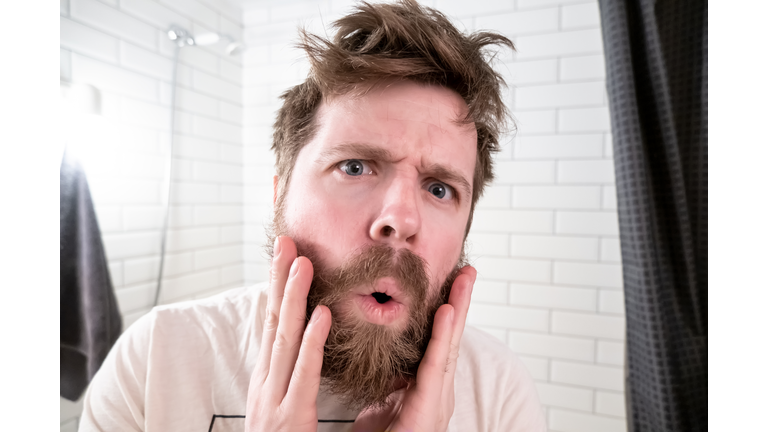 Overgrown man, with a shaggy hairstyle and beard, sees himself reflected in the mirror, he is shocked by his appearance.