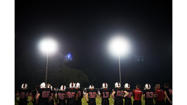 Rear view of American football players standing on illuminated field against sky