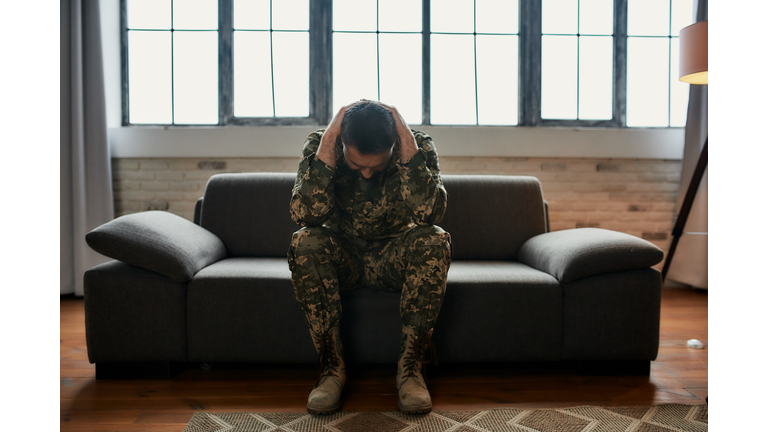 Constant pain. Full length shot of middle aged military man holding his head in pain and depression sitting on the couch. Soldier suffering from psychological trauma. PTSD concept