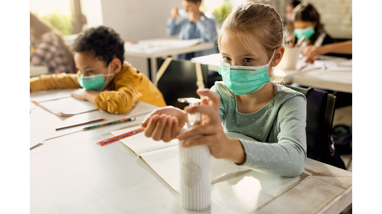 Elementary student wearing protective face mask and disinfecting her hands in the classroom.