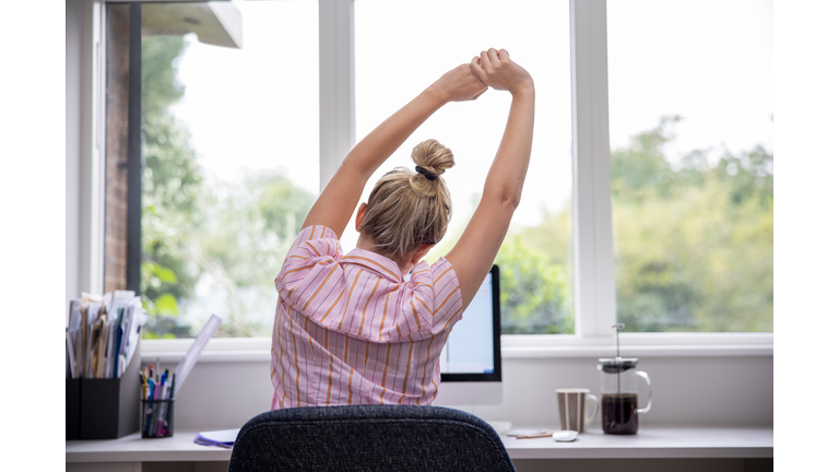 Rear View Of Woman Working From Home On Computer In Home Office Stretching At Desk