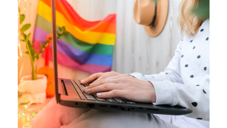Young millennial hippie woman sitting on balcony using laptop. LGBTQ rainbow flag on background. Online education tuition. Work from home. Modern office
