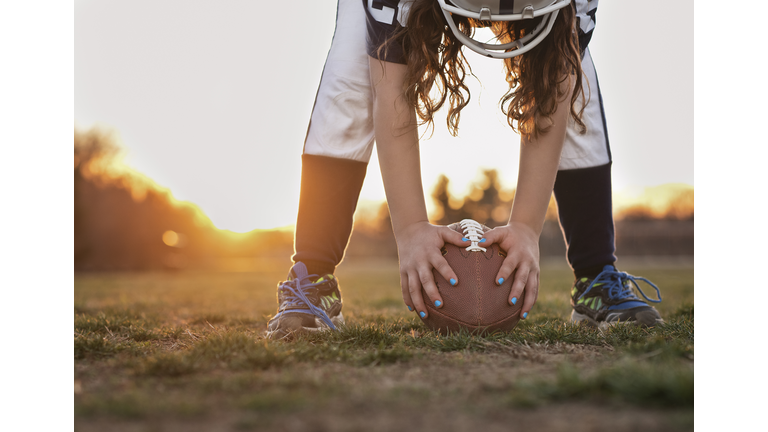 Low section of girl holding American football while standing on grassy field against sky during sunset