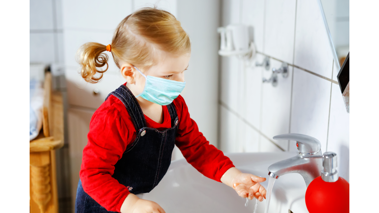 Toddler girl wear medical mask and cleaning, washing hands with soap and water. Child learning cleaning body parts during corona virus pandemic lockdown. Ppreventive measure and covid protection.