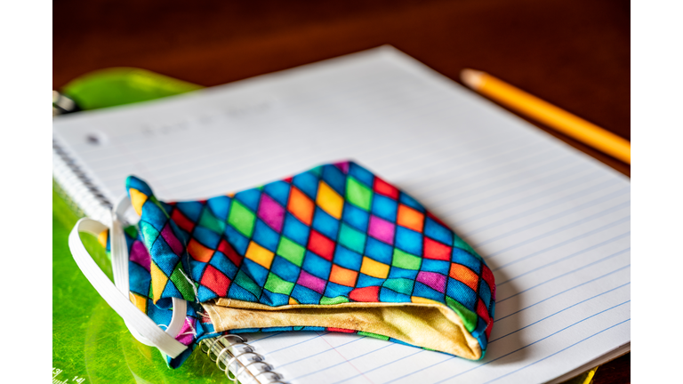 Face mask resting on top of back to school supplies of paper, pencil and a clip board