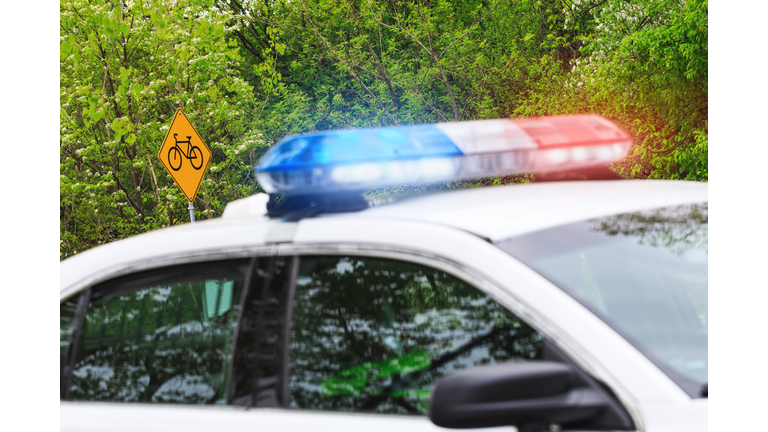 Bicycle or bike road sign with selective focus & police car with activated siren lights in blurred plan. A police patrol supervises the road reserved for cyclists or bicyclers.