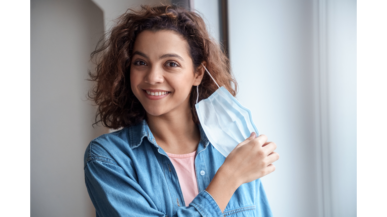 Happy Hispanic young woman takes off protective mask indoors.