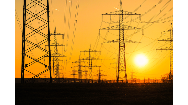 Electricity poles and electric power transmission lines against vibrant orange sky at sunset on a hot day with flickering air. High Voltage towers provide power supply over a long distance.