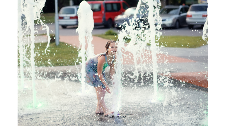 Little cute funny girl child bathes on a hot summer day in a public city fountain. The child runs, plays among the jets and splashes of water