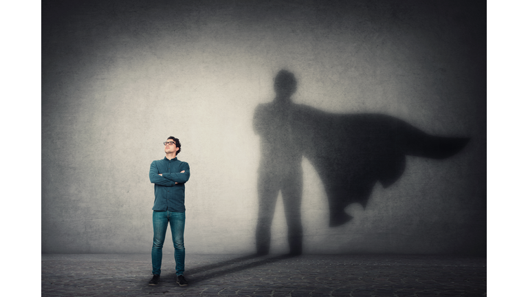 Brave man keeps arms crossed, looks confident, casting a superhero with cape shadow on the wall. Ambition and business success concept. Leadership hero power, motivation and inner strength symbol.