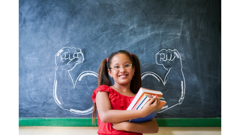 Hispanic Girl Holding Books In Classroom And Smiling