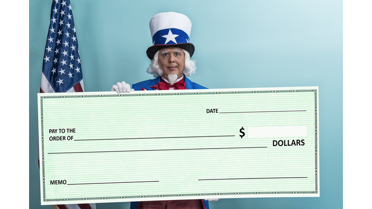 Uncle Sam holding a large check while standing against a teal wall, Concept - Tax cuts/tax refunds