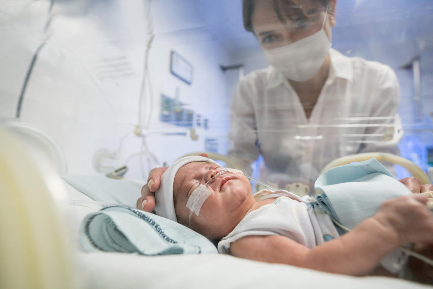 Loving mother looking at her newborn in an incubator while wearing a facemask