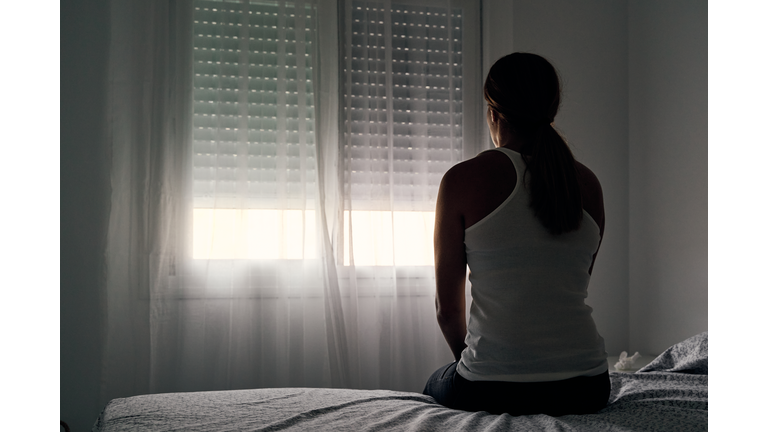 Rear view of an unrecognizable abused woman sitting on her bed looking out the window.