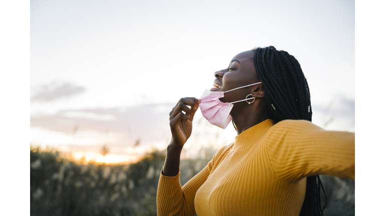 Happy teenage girl with eyes closed removing mask and inhaling in field during sunset