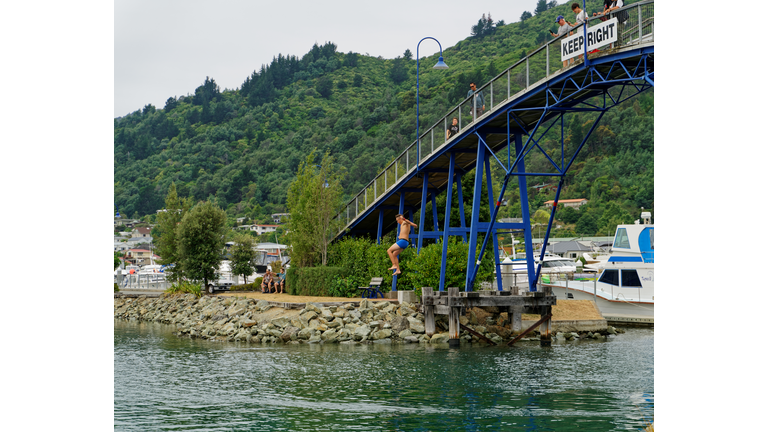 Young man jumps off the Marina footbridge into the sea beneath, while onlookers film the jump. Picton Marina, Marlborough Sounds, New Zealand.