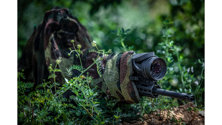 Sniper in camouflage suit in an ambush among the bushes. The shooter aims from a carbine with an optical sight. Sniper in position.