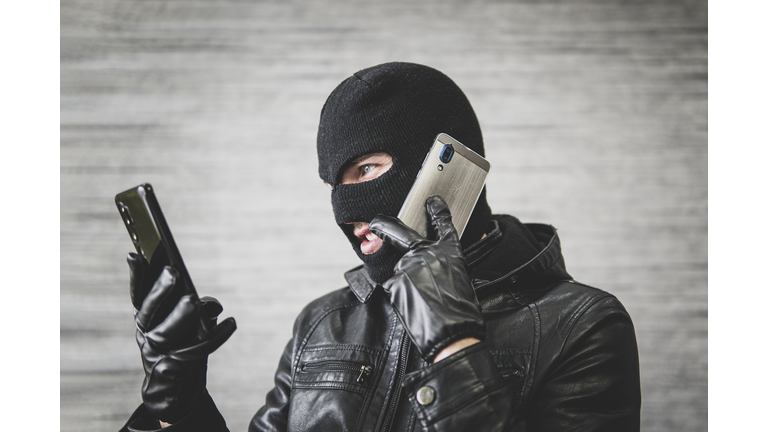 Young adult in black clothes with hidden face. Ill-intended fraudster uses mobile. Fraudster calls. Scam. Mobile racket. Hacker hijacks by phone. Cellphone account fraud.