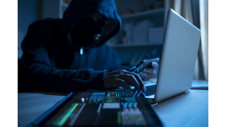 Anonymous person in the hood sitting in front of computer working with laptop and mobile phone