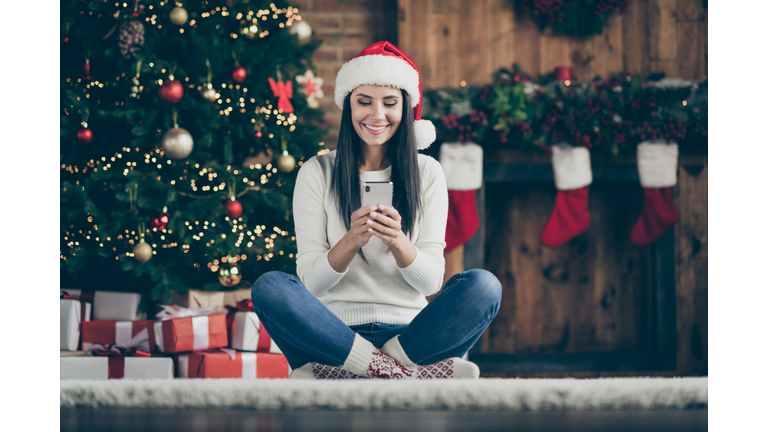 Full length photo of positive girl sir comfort floor carpet use her smartphone type text messages search christmas discounts wear santa claus hat in house with x-mas decoration indoors