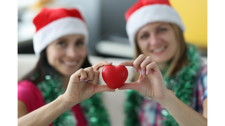 Two smiling woman in santa hats hold heart.