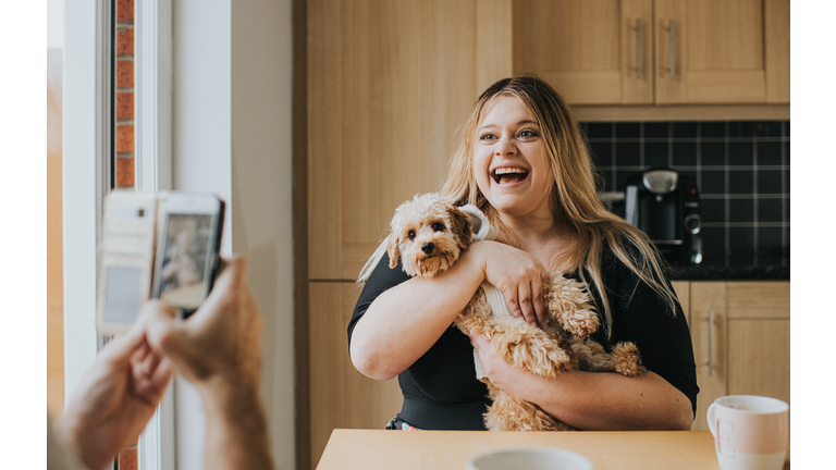 Woman cradles a Dog while Man photographs them in a domestic environment.