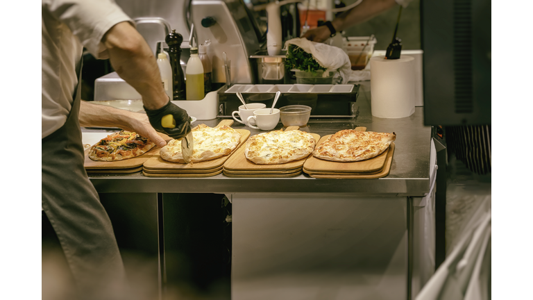 Pizzeria kitchen. Chef cook in apron and gloves cuts freshly baked pizza with roller knife.