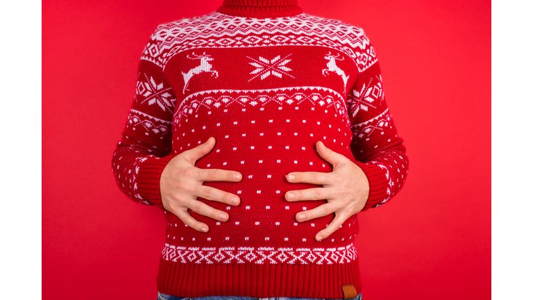 Holidays overeating concept. Close up photo of men in red winter sweater with deers holding his big tummy isolated on red background