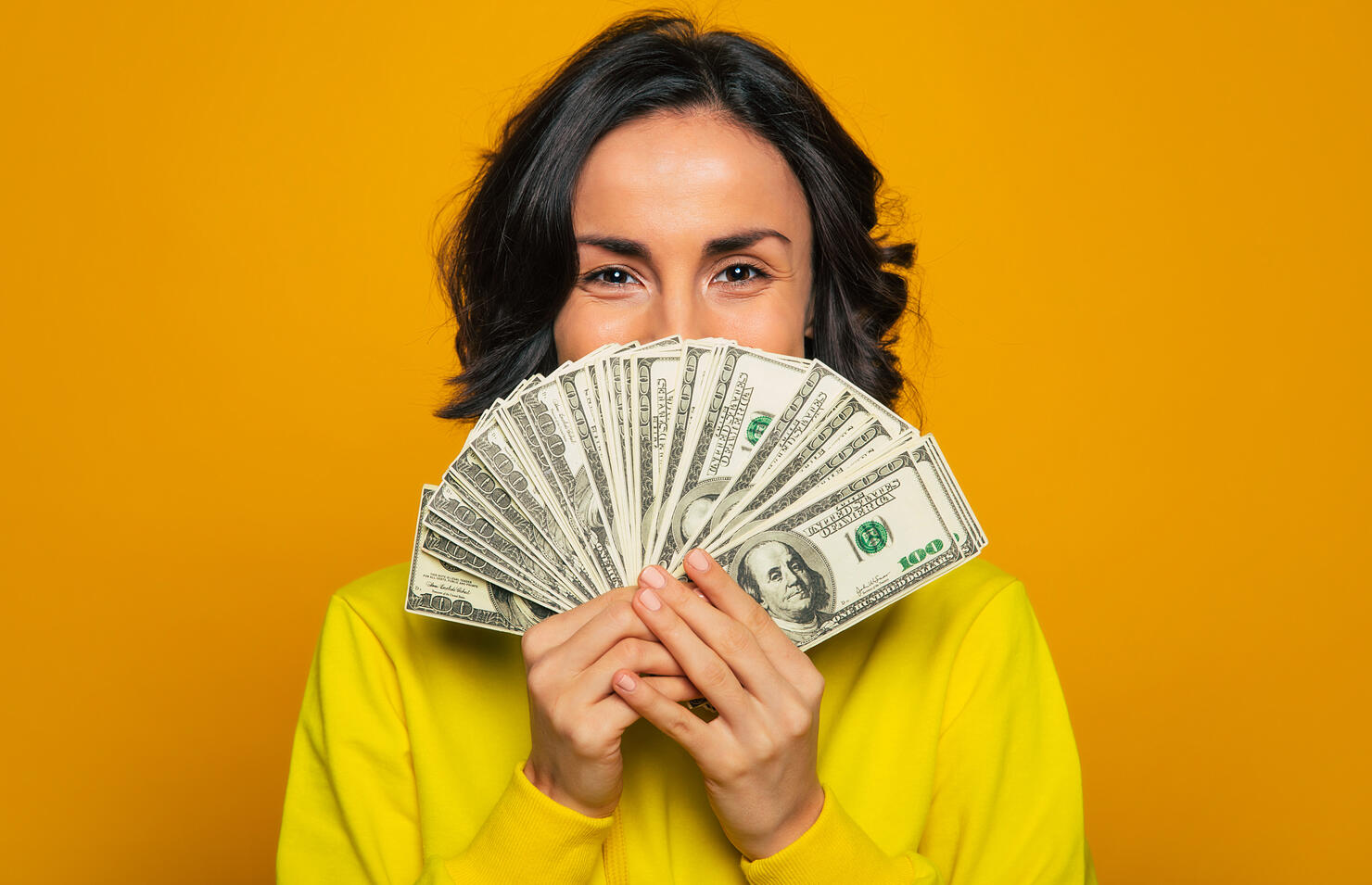 Here is my salary! Close-up photo of a young girl in a yellow hoodie, smiling with  her eyes, hiding her face behind big amount of money in her hands.