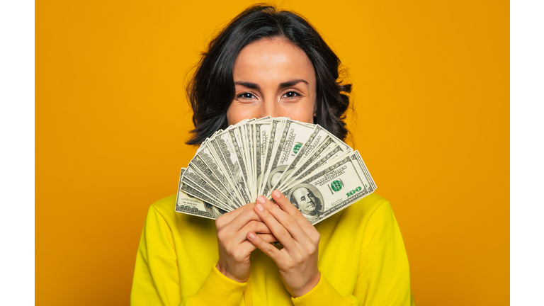 Here is my salary! Close-up photo of a young girl in a yellow hoodie, smiling with  her eyes, hiding her face behind big amount of money in her hands.