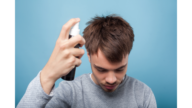 Portrait of young man applying spray to his hair. indoor studio shot isolated on blue background