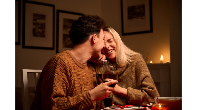 Happy young couple in love hugging, laughing, drinking wine, enjoying talking, having fun together celebrating Valentines day dining at home, having romantic dinner date with candles sitting at table.