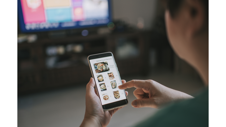 Online food delivery mobile app shown on smart phone screen hold by asian woman hands in living room